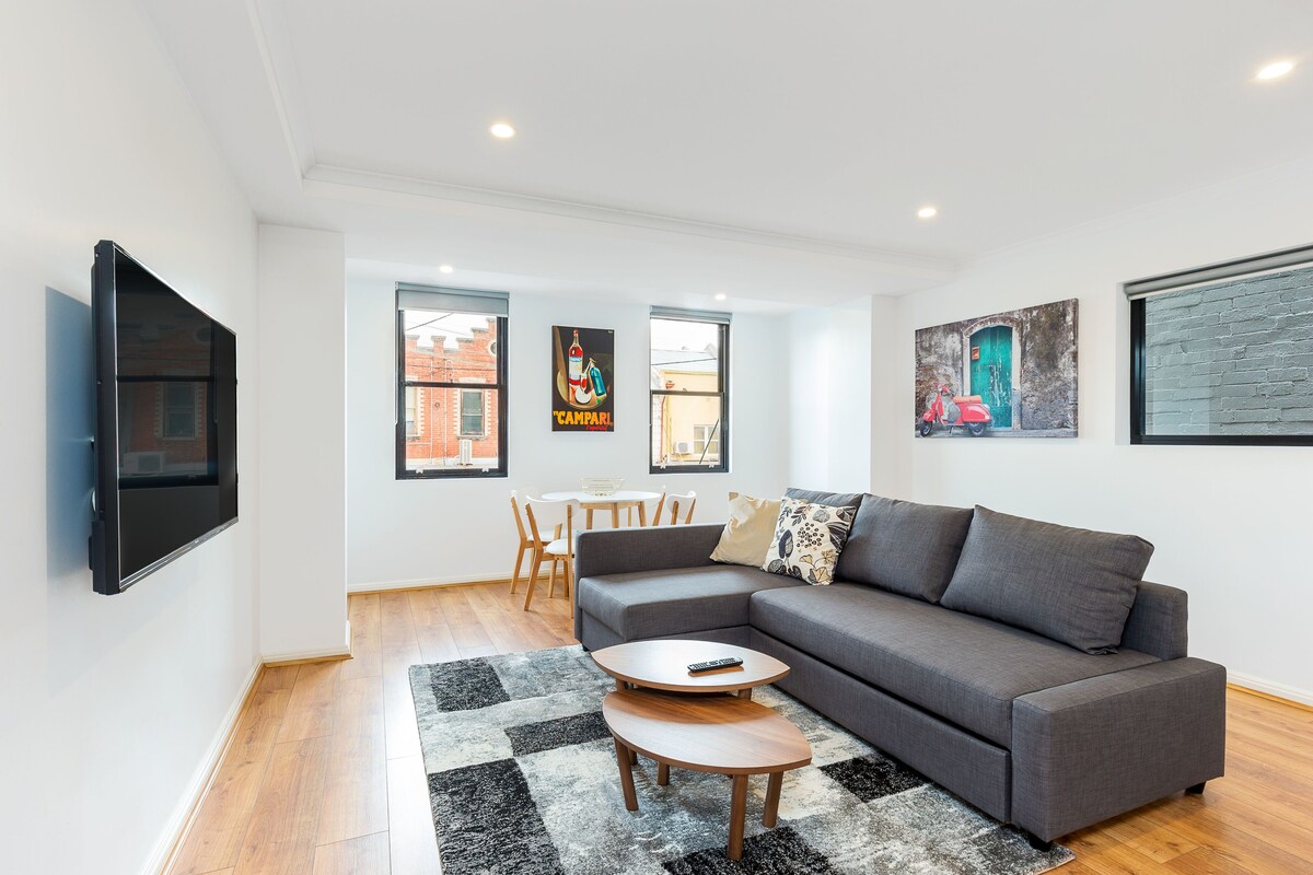 A modern living room with a grey sofa, a coffee table, and a flat-screen TV mounted on the wall. There are two windows with a view of buildings outside.