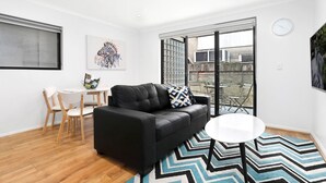 A modern living room with a black leather sofa, a round white table, and a zigzag patterned rug. There is a dining area with a small table and chairs, and a large window providing a view of the outside. - Balmain Modern Apartments (Rozelle)