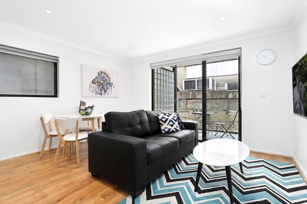 A modern living room with a black leather sofa, a round white table, and a zigzag patterned rug. There is a dining area with a small table and chairs, and a large window providing a view of the outside.