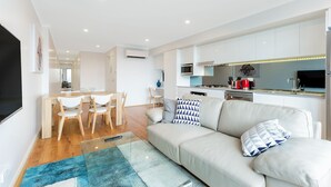 A modern living room with a sofa, a coffee table, and a TV. There is a dining area with chairs and a table. The kitchen is visible with cabinets and appliances. - Balmain Modern Apartments (Rozelle)