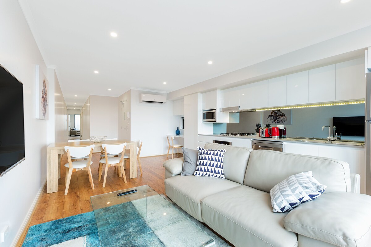A modern living room with a sofa, a coffee table, and a TV. There is a dining area with chairs and a table. The kitchen is visible with cabinets and appliances.