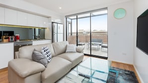 A modern living room with a sofa, a glass coffee table, and a sliding glass door leading to an outdoor patio. - Balmain Modern Apartments (Rozelle)