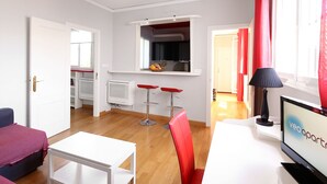 Uma sala de estar moderna com um sofá branco, uma mesinha branca e uma cadeira vermelha. Há uma televisão em um suporte, um abajur e uma janela com cortinas vermelhas.