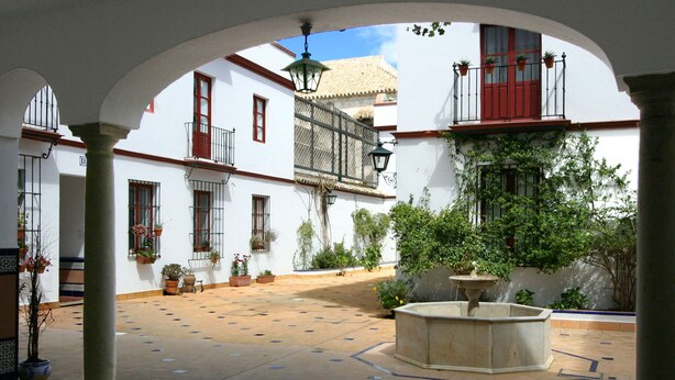 Um pátio aberto com uma fonte, plantas em vasos e edifícios brancos com molduras e varandas vermelhas.