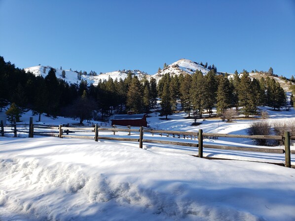 Un paysage enneigé avec une clôture en bois, un bâtiment rouge et une chaîne de montagnes en arrière-plan.