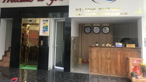 Un hall d'accueil d'hôtel avec une pancarte indiquant « Bienvenue », un comptoir de réception, des horloges affichant les heures des fuseaux horaires du Vietnam, de la Corée et de l'Angleterre, et un comptoir de réception en bois sur lequel est inscrit le mot « Lê Tân ».
