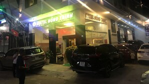 L’entrée d’un hôtel, avec une enseigne indiquant « HÔTEL HONG THINH » et un drapeau. Des voitures sont garées devant, et une personne se tient près de l’entrée.