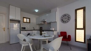 A modern kitchen with white cabinets, a round dining table with white chairs, a red chair, and a wall clock. - Mare Verde 1 (Costa Adeje)