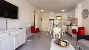 A modern living room with a flat-screen TV, a dining area with a white table and red chairs, a kitchen with white cabinets, and a clock on the wall. - Mare Verde 1 (Costa Adeje)