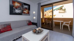 A modern living room with a grey sofa, a white coffee table, and a large abstract painting. There is a sliding glass door leading to an outdoor area with a white table and chairs. - Mare Verde 1 (Costa Adeje)