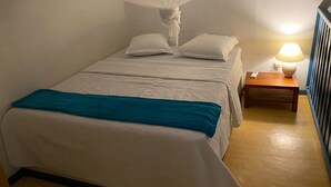 En pænt redt seng med et hvidt og teal sengetæppe, en hvid himmel og et natbord med en lampe.