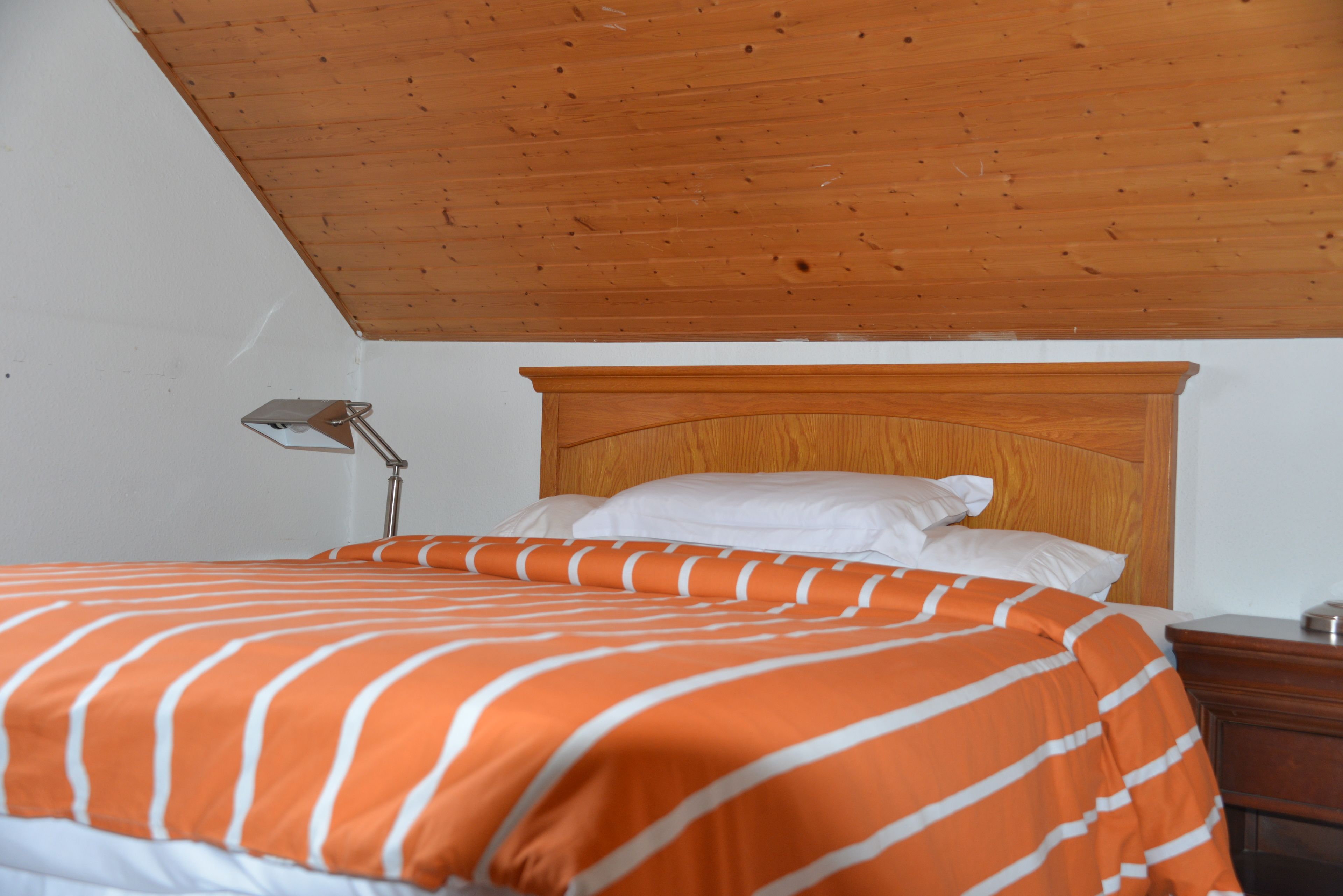 Habitación de hotel con techo de madera, cama con colcha a rayas naranja y blancas, almohadas blancas y mesita de noche con lámpara.