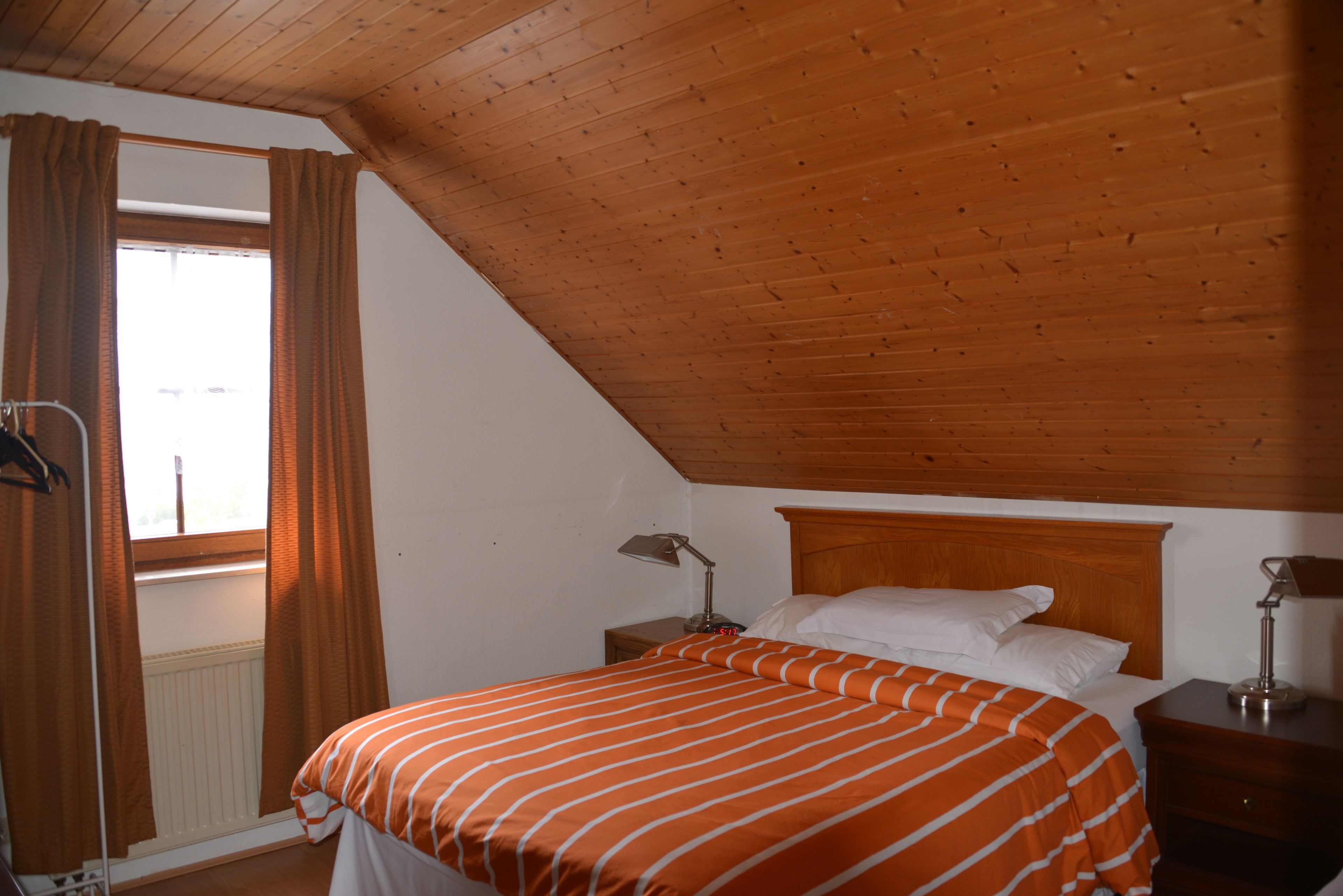 A bedroom with a sloped ceiling, a bed with an orange-striped bedspread, two bedside lamps, and a window with brown curtains.