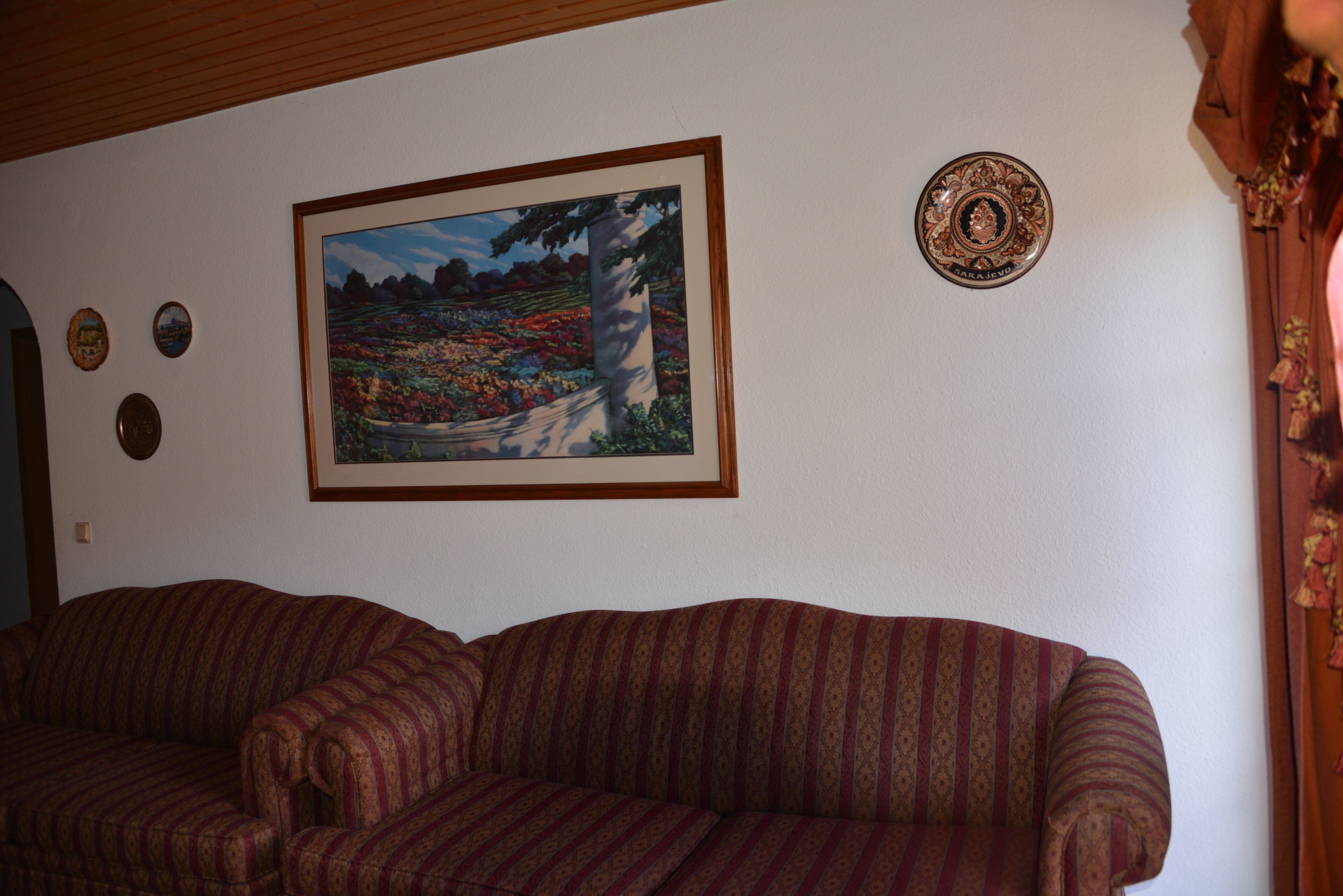 Un salon avec un canapé à motifs, un tableau représentant un paysage encadré et des assiettes décoratives accrochées au mur.