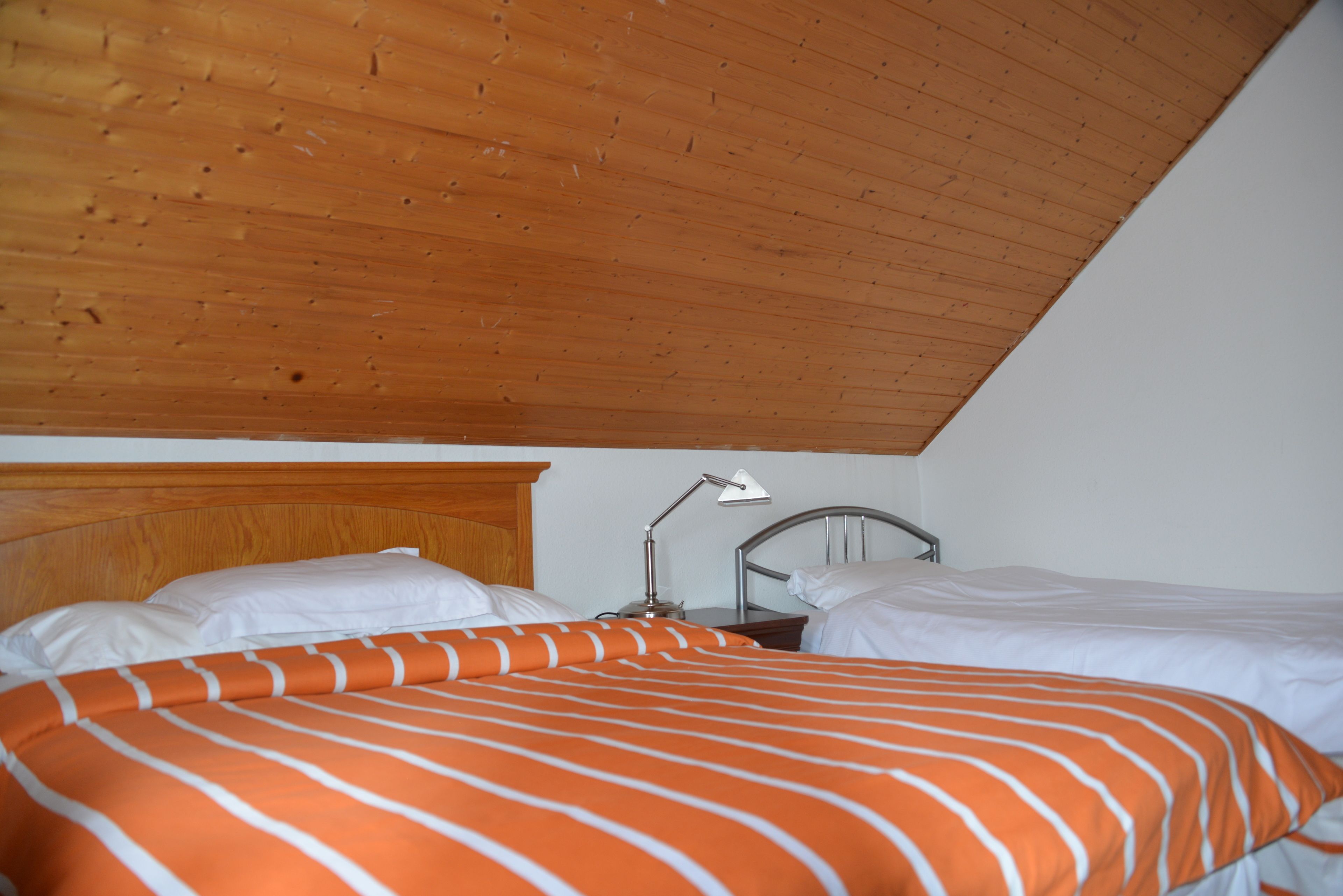 Ein Schlafzimmer mit schrägen Wänden, einem Holz-Kopfteil, zwei Betten mit weißer Bettwäsche und einem Nachttisch mit Lampe.