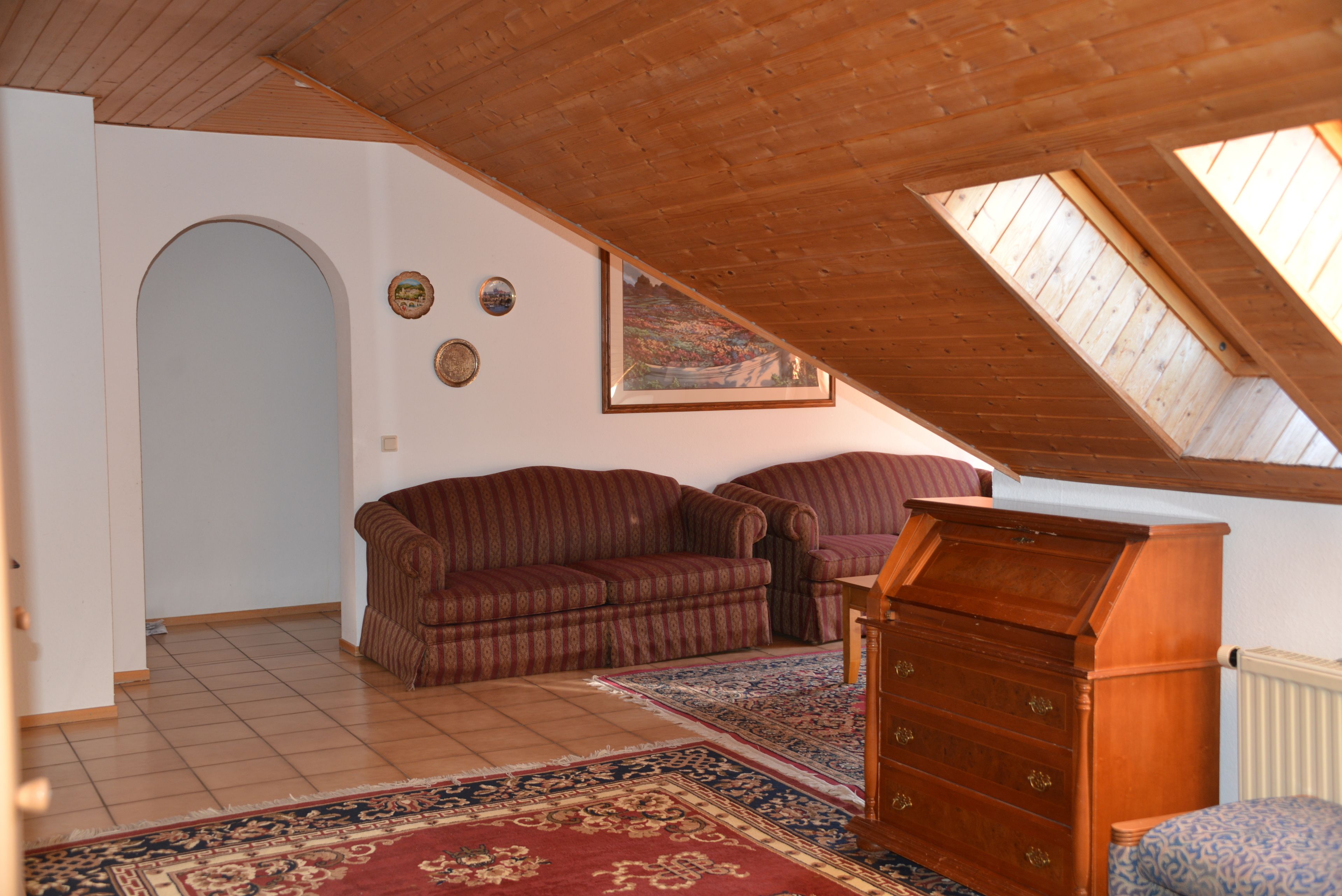 Una habitación con techo inclinado, un aparador de madera, un tapete estampado, un sofá y un cuadro en la pared.