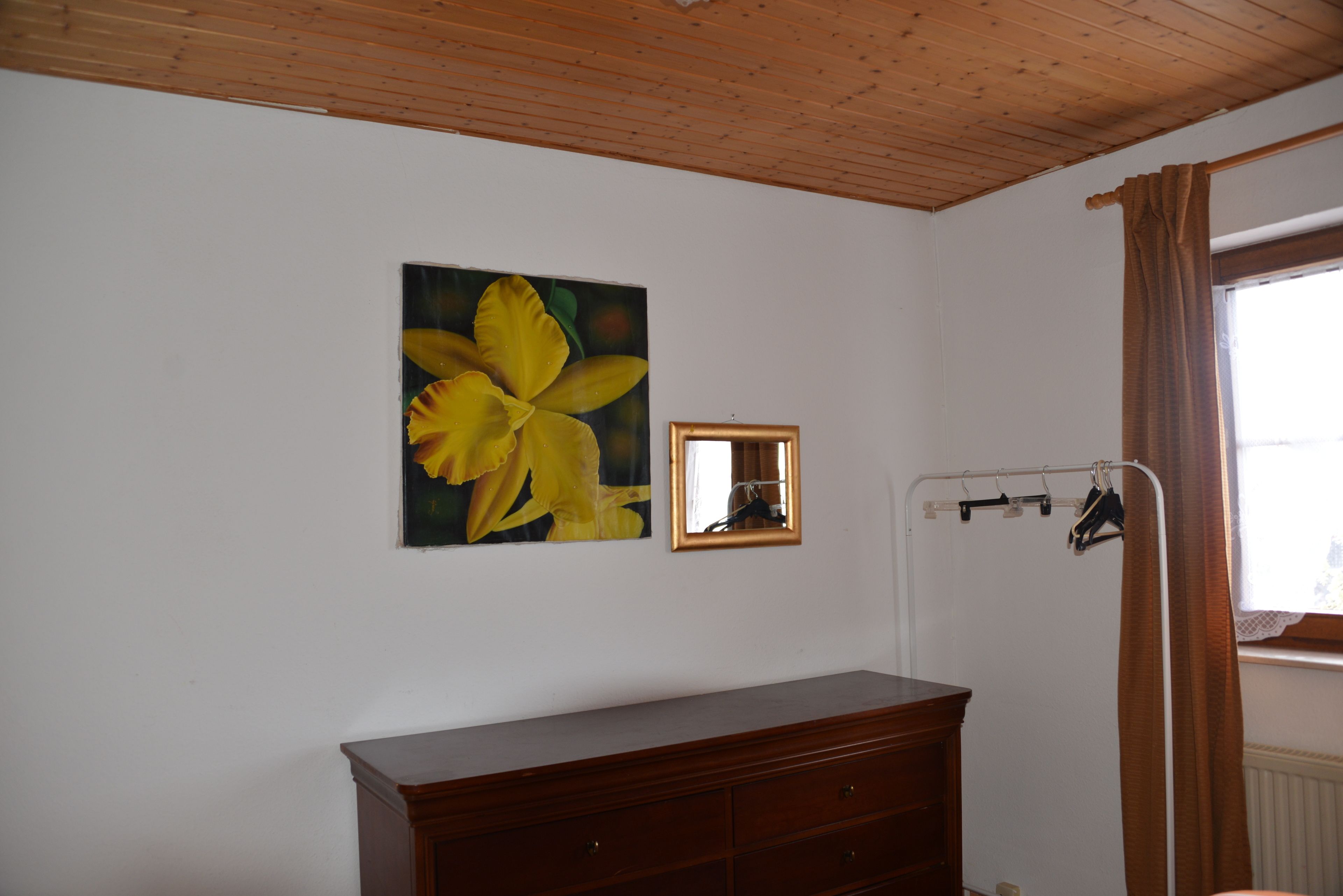 A room with a wooden ceiling, a large yellow flower painting, a mirror, a wooden dresser, and a window with curtains.