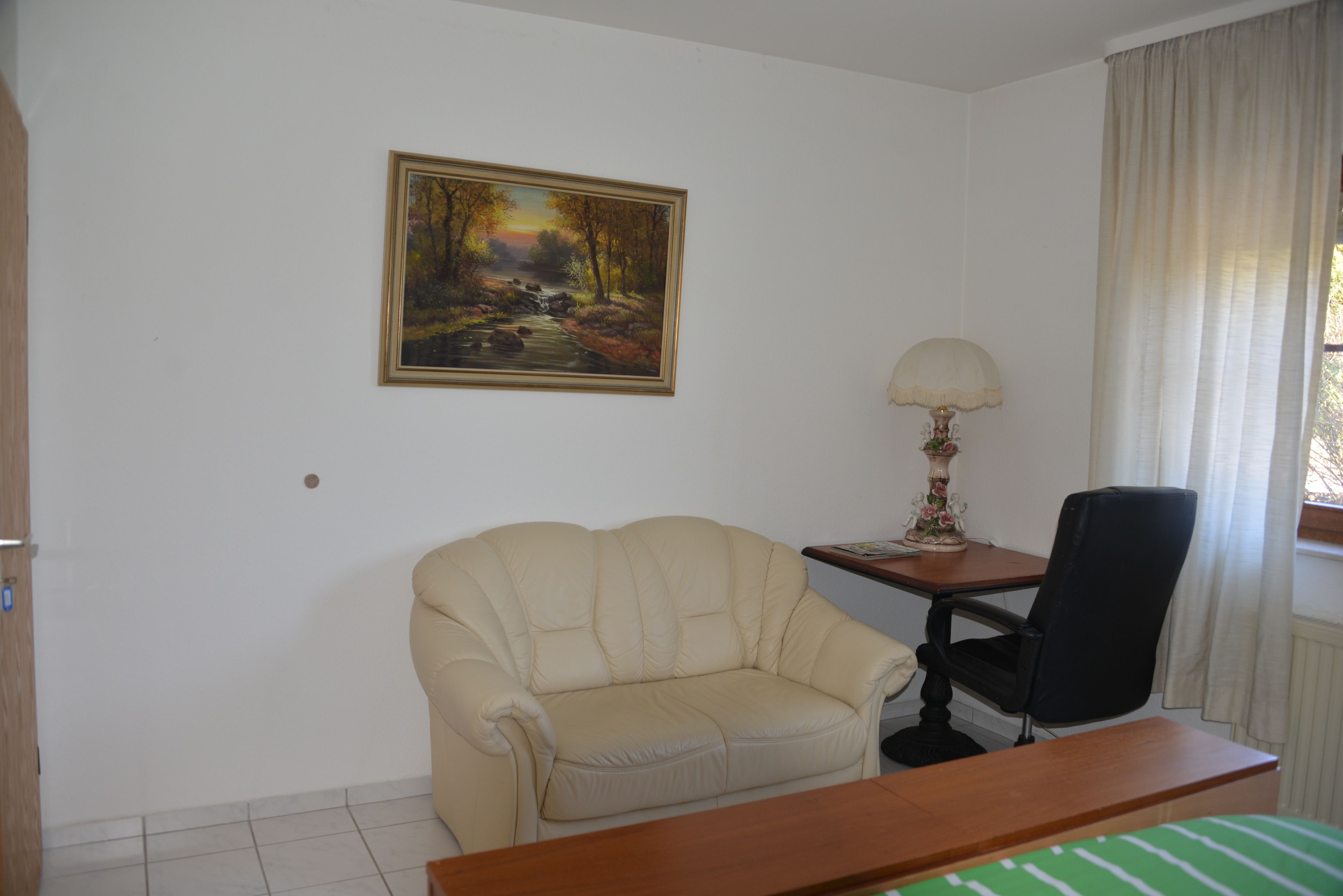 Ein Zimmer mit einem beigefarbenen Sofa, einem Holztisch, einem schwarzen Bürostuhl, einer Lampe und einem Landschaftsbild an der Wand.