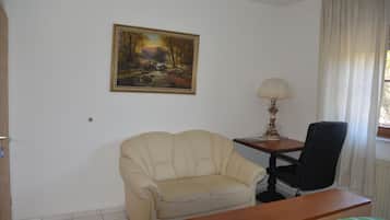 Ein Zimmer mit einem beigefarbenen Sofa, einem Holztisch, einem schwarzen Bürostuhl, einer Lampe und einem Landschaftsbild an der Wand.
