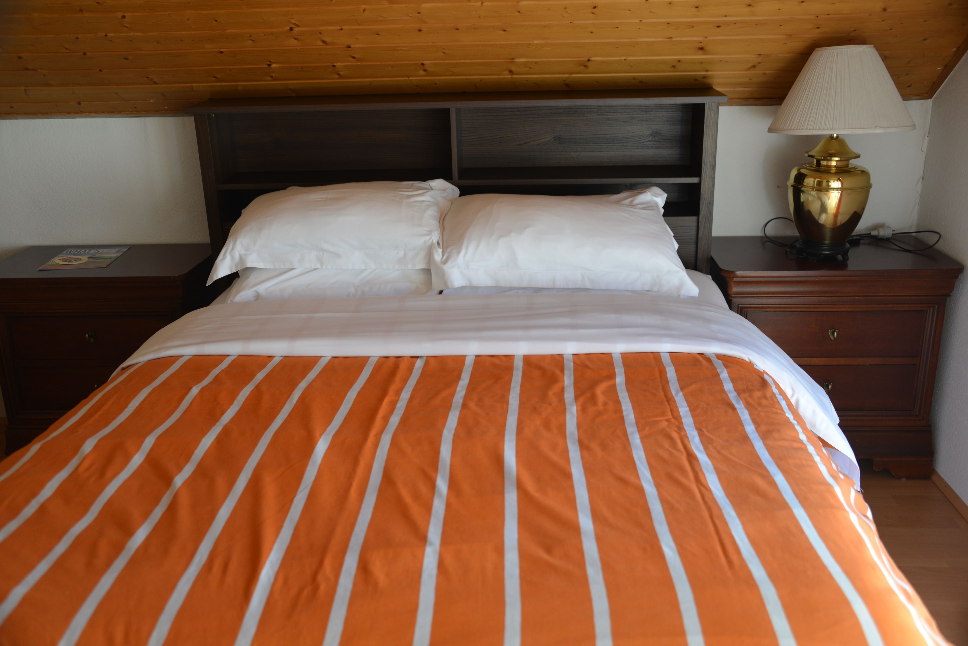 Una cama con colcha a rayas anaranjada y blanca, cabecera de madera, mesita de noche con lámpara y cómoda.