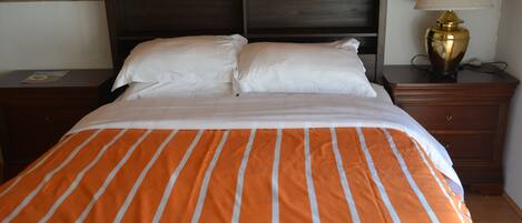 A bed with an orange and white striped bedspread, a wooden headboard, a nightstand with a lamp, and a drawer unit.