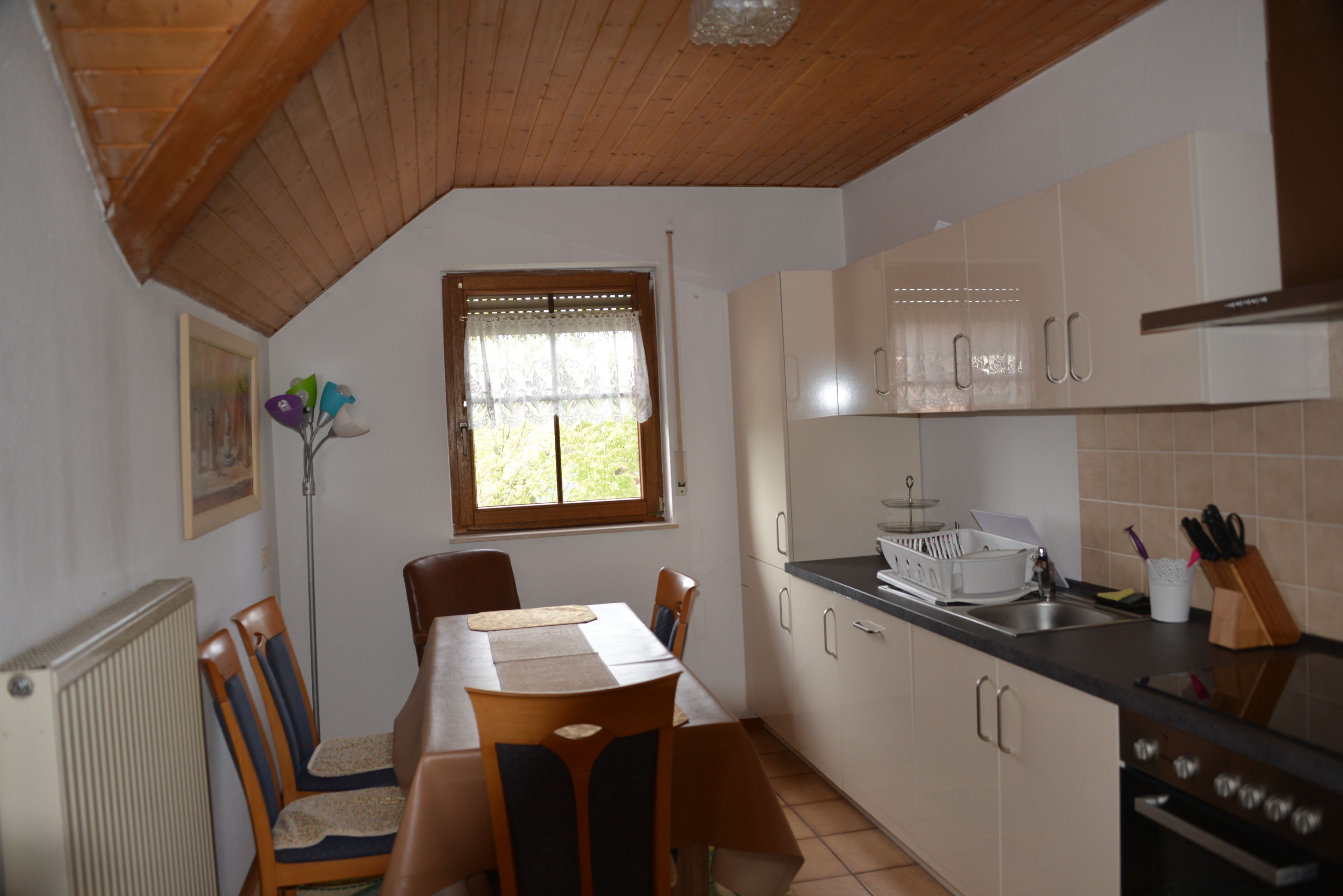 Una cocina con armarios de madera, una mesa de comedor con sillas, una ventana con cortinas de encaje y un radiador.