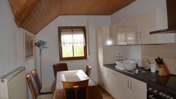 Una cocina con armarios de madera, una mesa de comedor con sillas, una ventana con cortinas de encaje y un radiador.