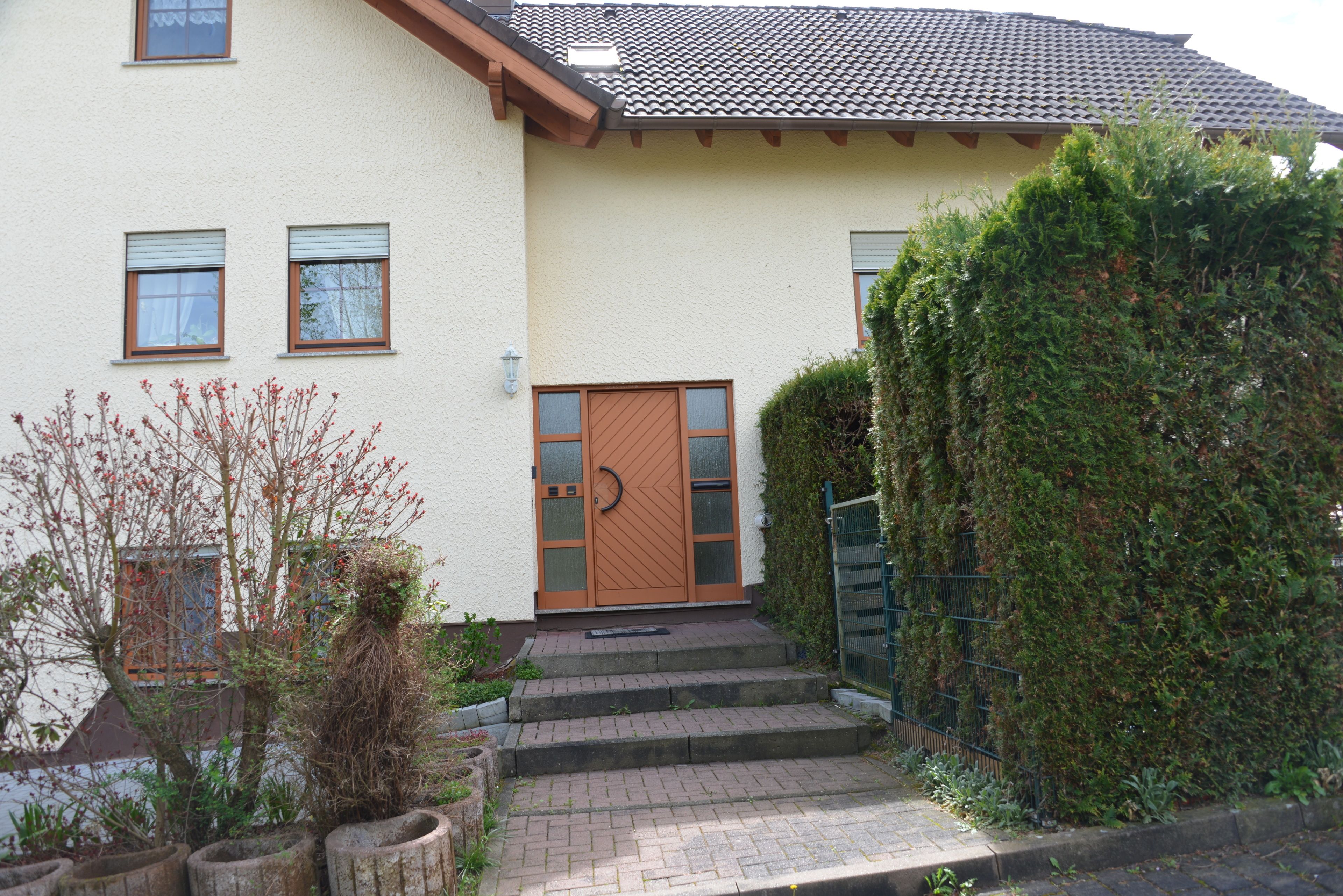 Ein zweigeschossiges Haus mit brauner Tür, einer kleinen Veranda und einem Garten mit Pflanzen und einem Baum.