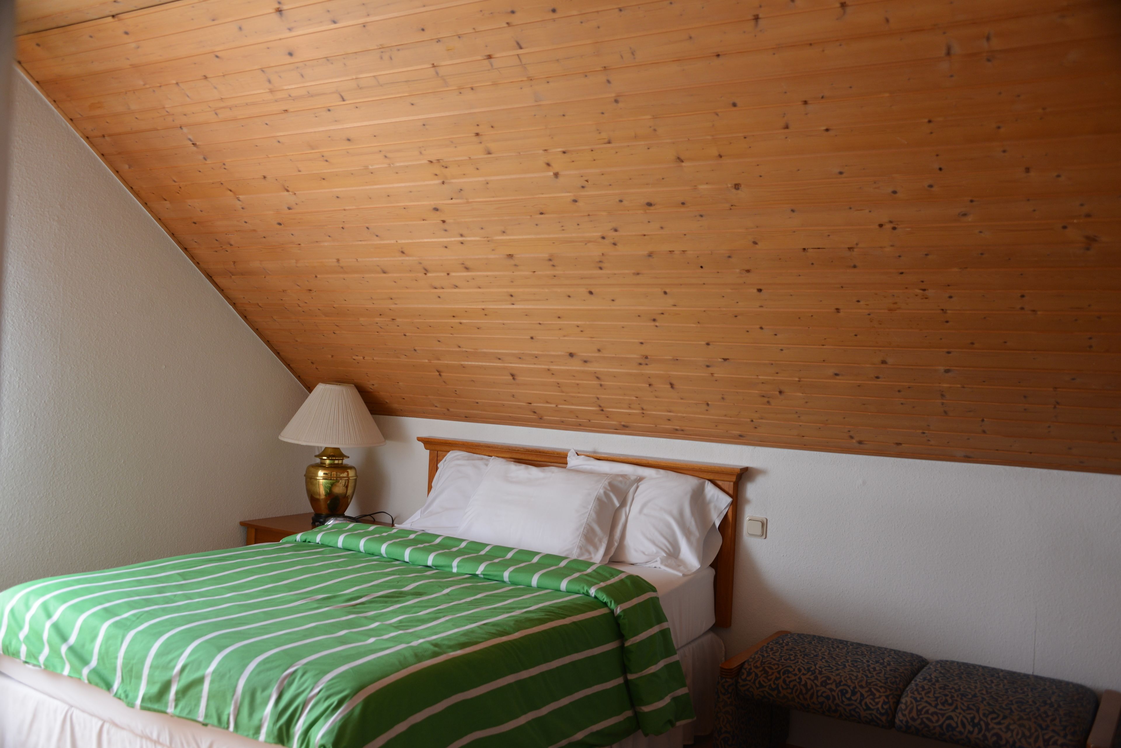 Une pièce avec un plafond en bois incliné, un lit recouvert d’une couvre-lit à rayures vertes, une lampe de chevet et un banc à motifs.