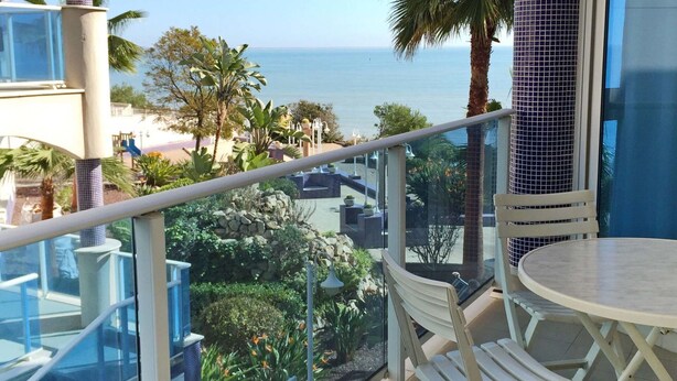 A balcony with a glass railing overlooking a beach and ocean. There is a white table and two white chairs. - Beautiful apartment for 4 people with pool, A/C, WIFI, TV and balcony (Ulldecona)