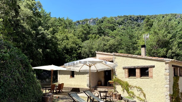 Un rústico edificio de piedra con mesas al aire libre y sombrillas, rodeado de exuberante vegetación y un paisaje montañoso.