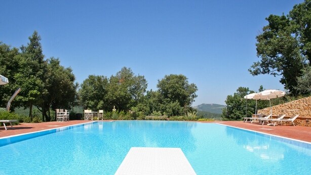 A clear blue swimming pool with a white deck in the center, surrounded by lounge chairs and umbrellas, with trees and a mountainous backdrop. - Apartment overlooking the Sienese hills, swimming pool and free wi-fi (Casole d'Elsa)