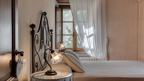 A bedroom with a bed, a bedside table with a lamp, a window with curtains, and a radiator. - Apartment overlooking the Sienese hills, swimming pool and free wi-fi (Casole d'Elsa)