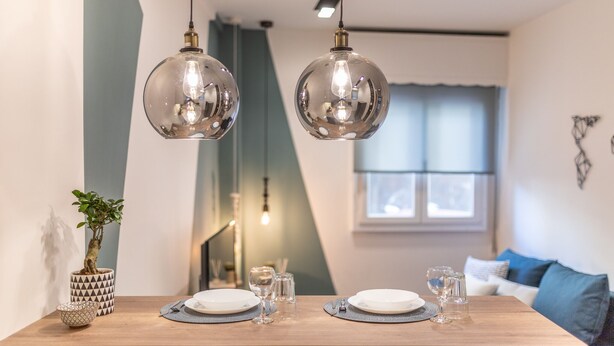 En matplats med ett träbord dukat för två personer, två hängande taklampor, en krukväxt och en modern väggdesign.