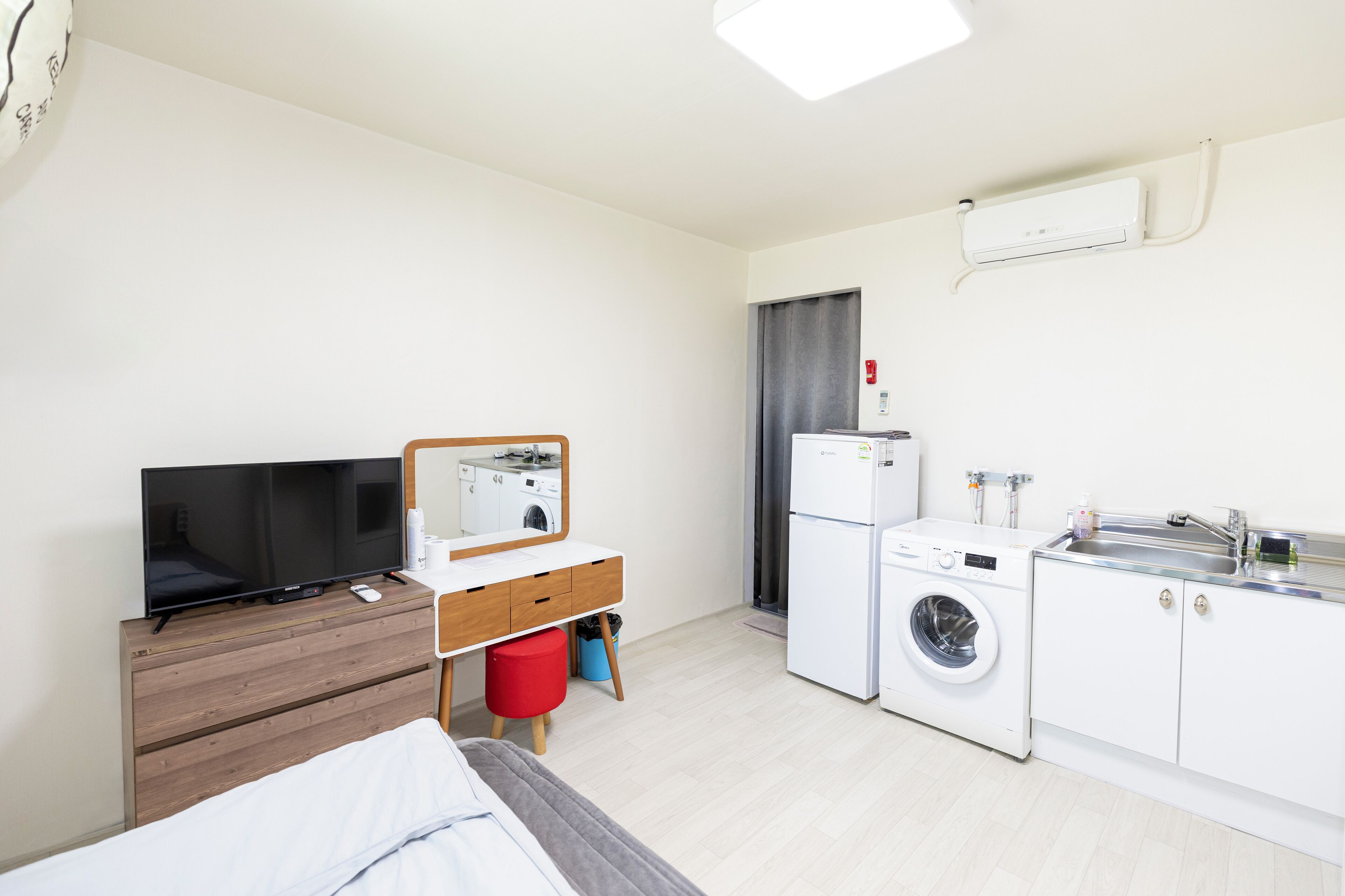 Ett kompakt hotellrum med en säng, en TV, ett litet skrivbord med en spegel, en tvättmaskin och ett handfat.