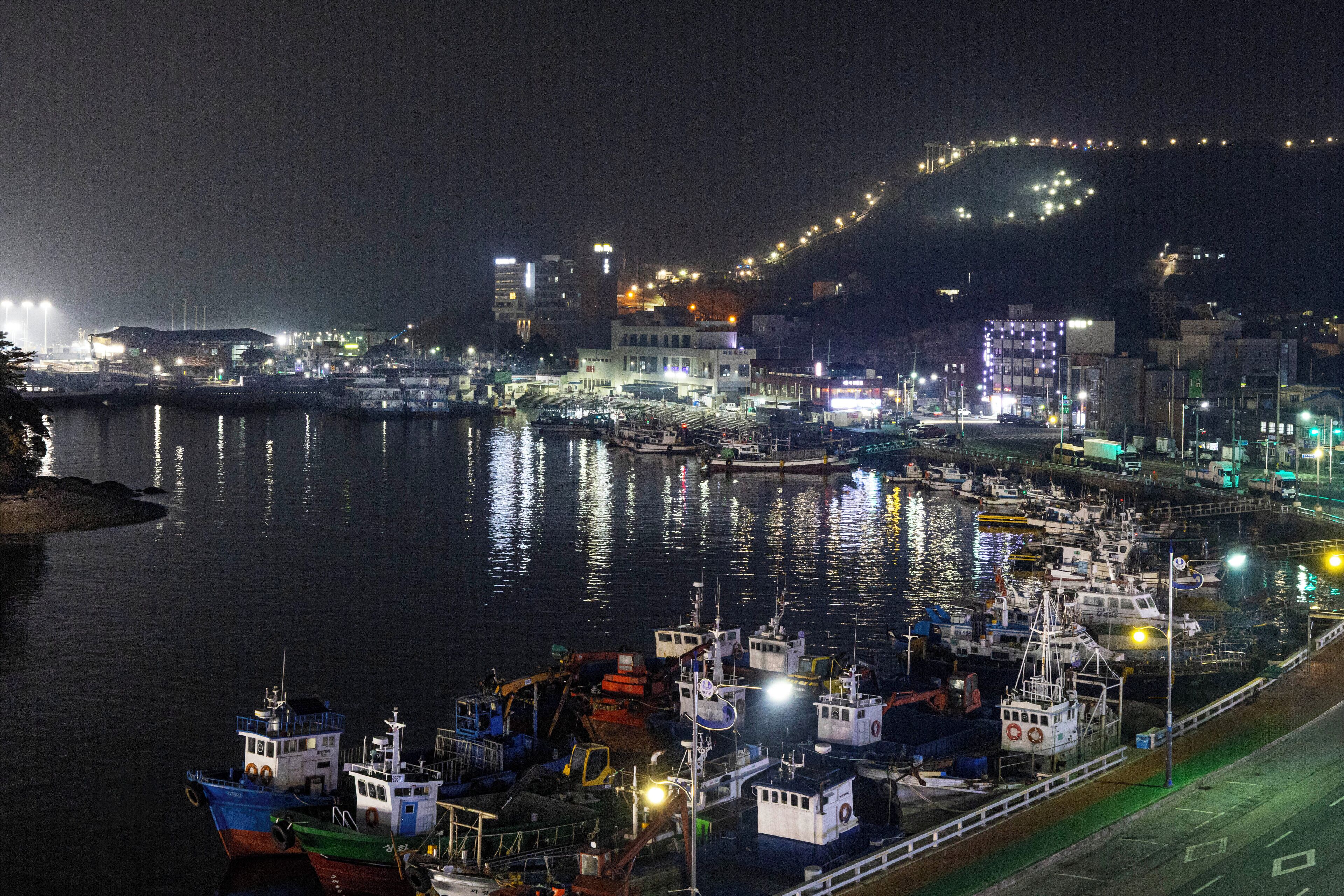 A nighttime marina with boats docked, a cityscape in the background, and a hill with lights.