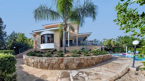 A modern house with a stone circular patio, a pool, and lush greenery. - Superb Villa in Coral Bay Paphos, with Half Cost of the HEATED POOL Nov-Mar 26 (Paphos (Pafos))