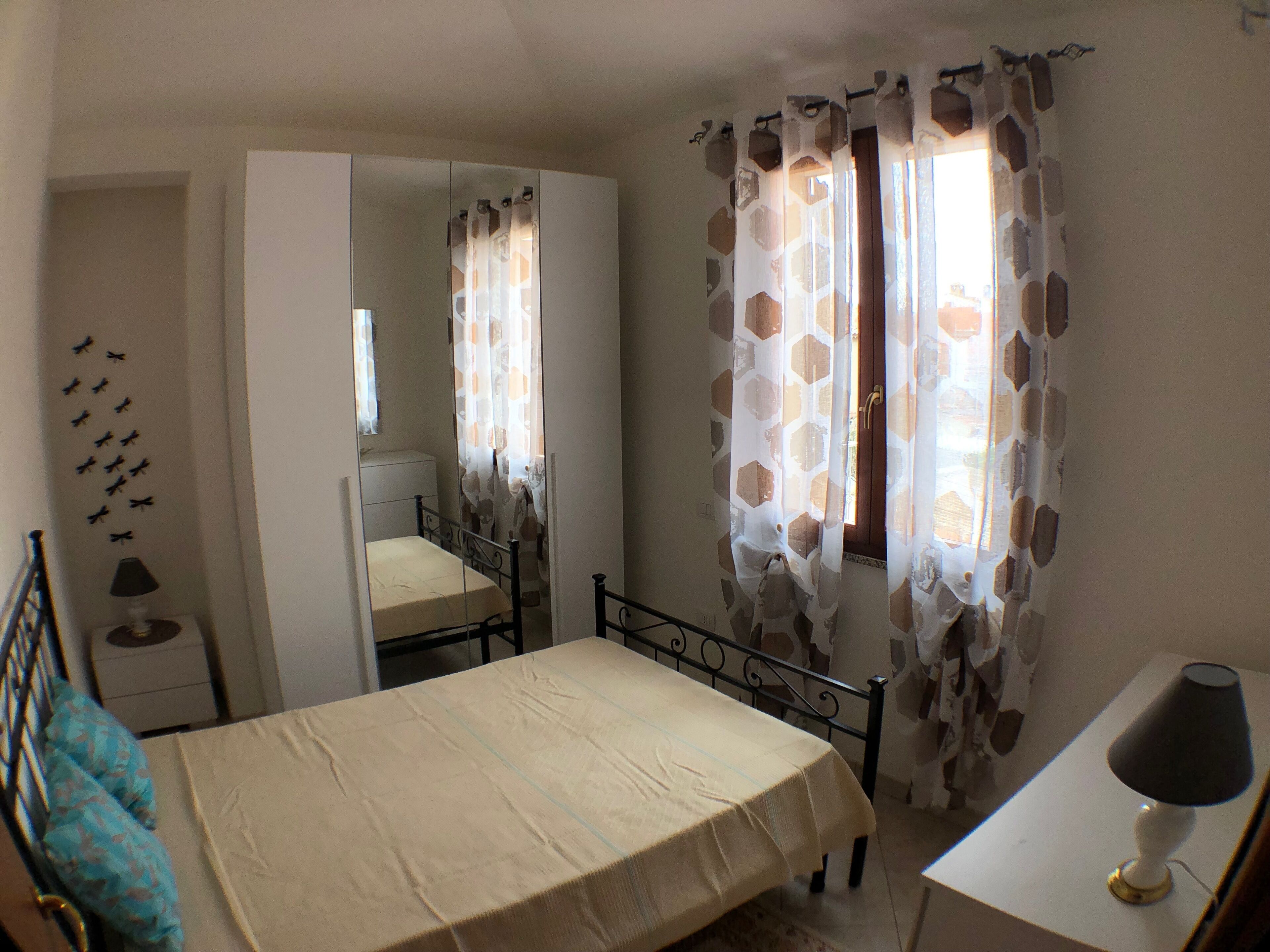 A bedroom with a bed, a bedside table, a lamp, a mirror, and a window with patterned curtains.