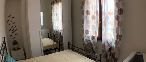A bedroom with a bed, a bedside table, a lamp, a mirror, and a window with patterned curtains.