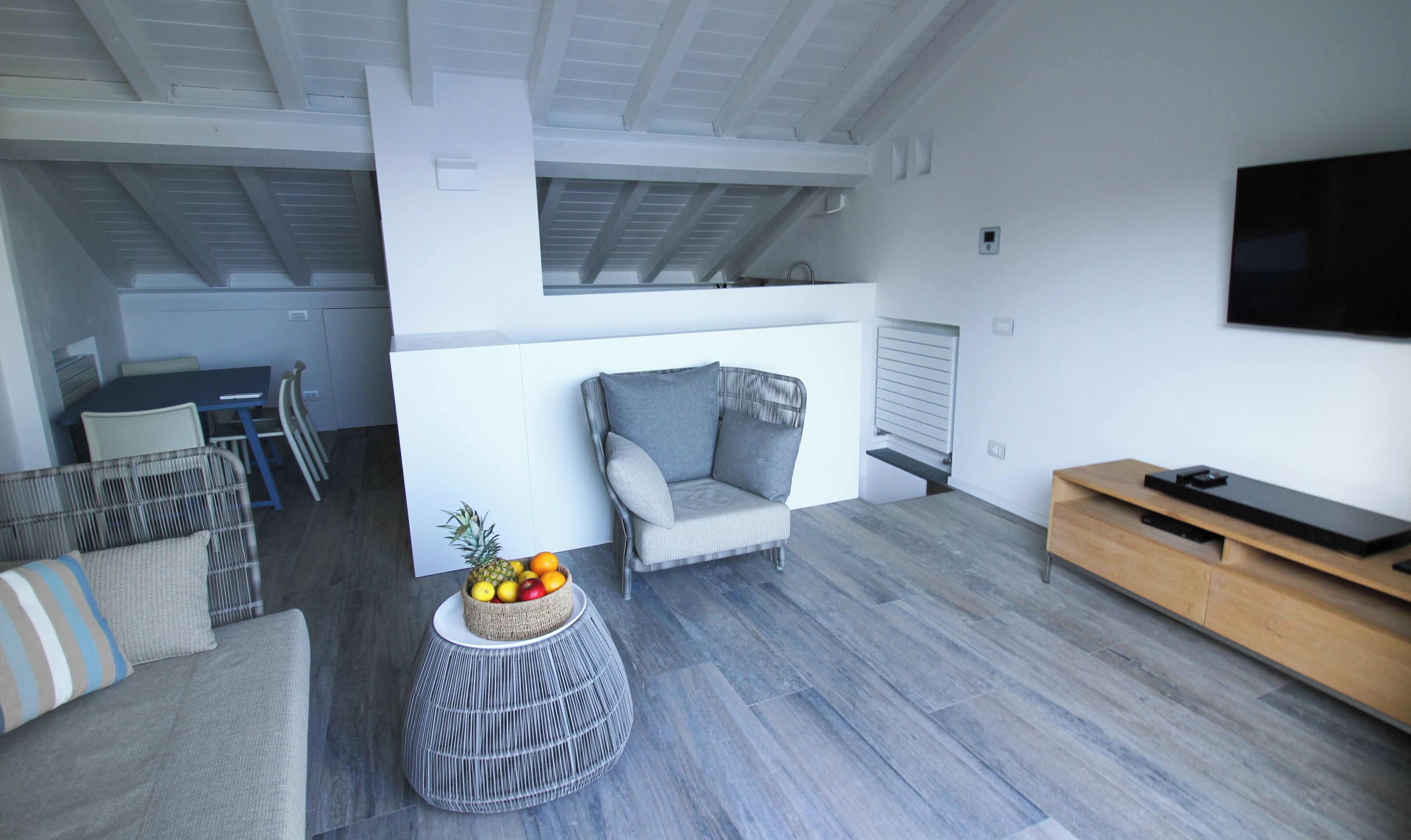 Un soggiorno moderno con un divano grigio, un poggiapiedi in vimini con della frutta, un mobile TV in legno e, sullo sfondo, un'area pranzo.