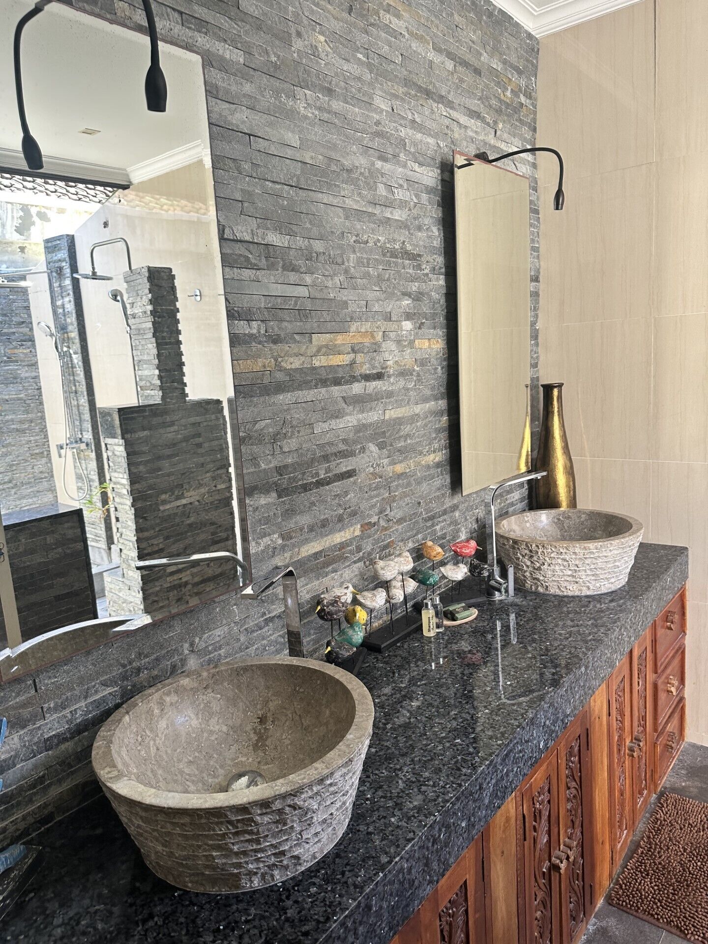 Une salle de bain avec un mur en pierre, un grand miroir, deux lavabos avec des cuves en pierre et un meuble en bois.