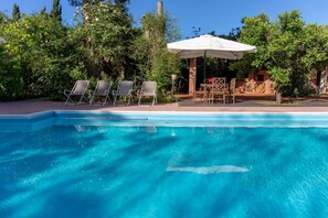 A swimming pool with clear blue water, surrounded by greenery and a wooden deck with lounge chairs and a table under a white umbrella. - Domus Sicily - Il Giardino di Michela - Heated Pool (Palermo)