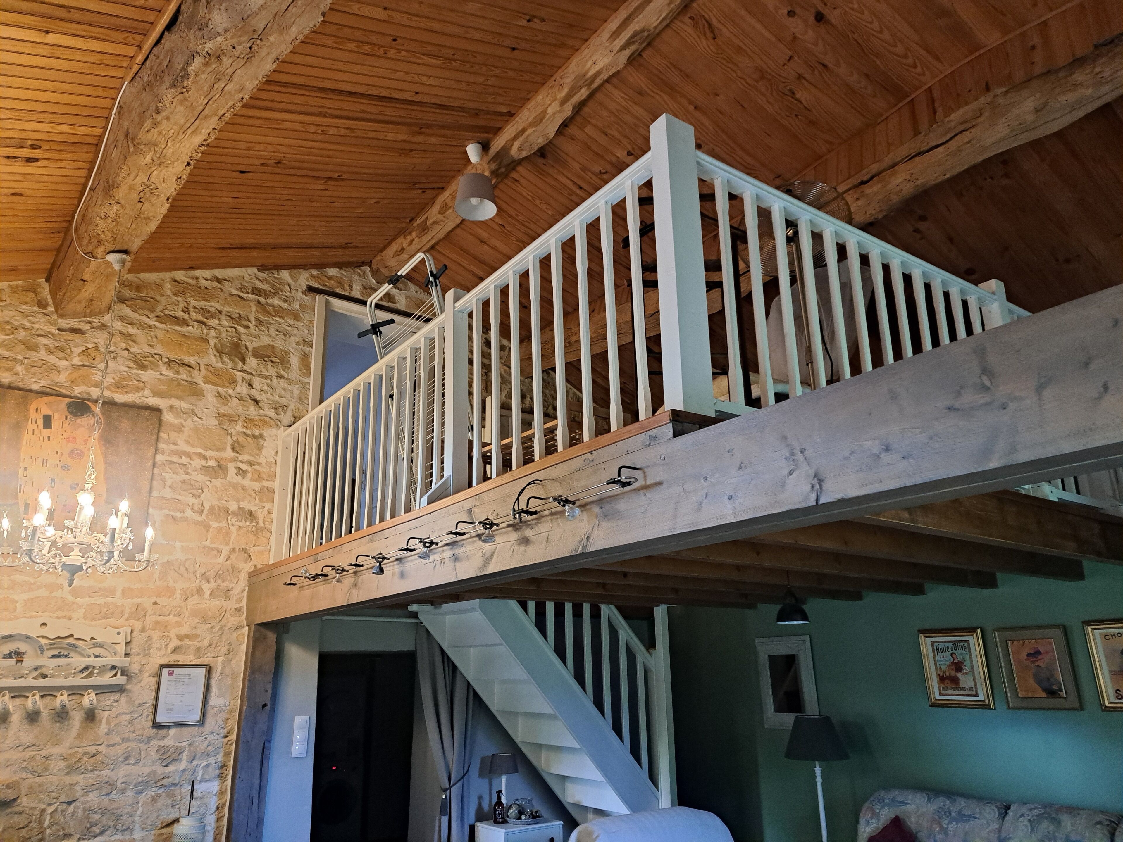Een rustieke kamer met zichtbare houten balken, een stenen muur, een balkon met witte hekwerk en een kroonluchter, plus een trap.