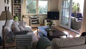 Ein Wohnzimmer mit Sofa, Sessel, Fernseher, Bücherregal und Blick durch eine Schiebetür.