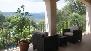 Ein Balkon mit Korbmöbeln, einem Blumentopf und Blick auf Grünflächen und Berge.