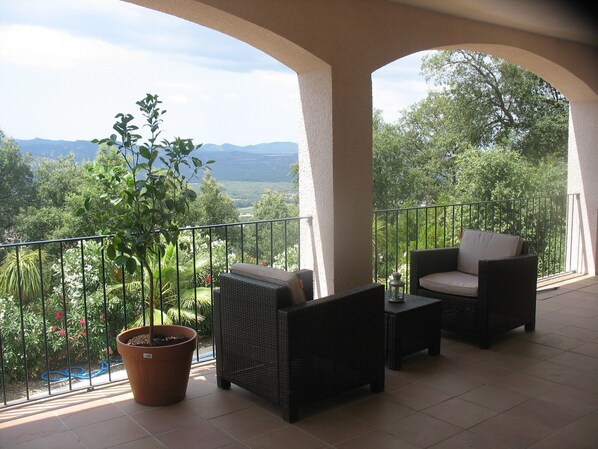 A balcony with wicker furniture, a potted plant, and a view of greenery and mountains.