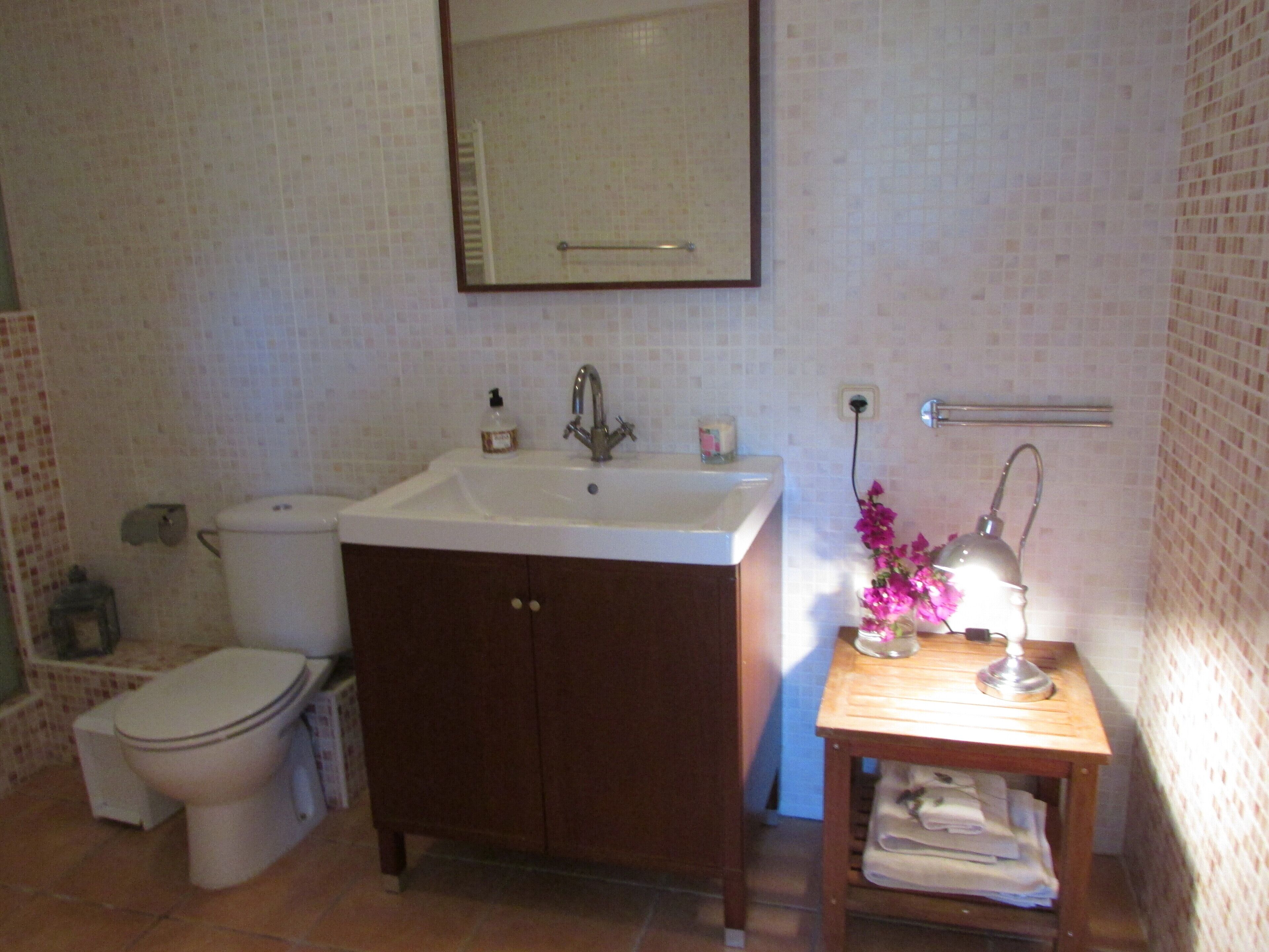 Une salle de bain avec un lavabo blanc, un miroir au-dessus, des toilettes et une petite table en bois sur laquelle se trouvent une lampe et des serviettes.