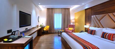 A hotel room with a large bed, a desk with a flat-screen TV, a chair, a desk lamp, and a window with curtains.