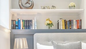 Een keurig opgemaakt slaapkamer met een bed, nachtkastje, lamp en een boekenplank met boeken en decoratieve objecten.