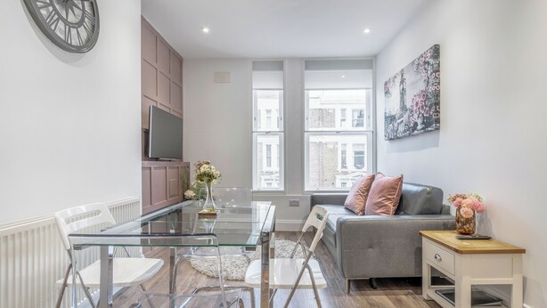 Een moderne woonkamer met een glazen eettafel, witte stoelen, een grijze bank en een klein houten bijzettafeltje met een vaas.